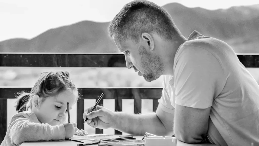 A child sitting at a table carefully coloring a printed page using crayons. The activity shows focus, creativity, and enjoyment during a quiet indoor learning moment.